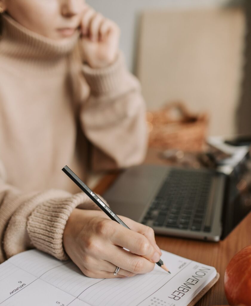 Close-up of a woman writing in a planner at home with a laptop and pumpkins.
